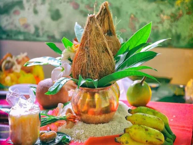 A traditional Hindu Kalash setup with a coconut placed on a copper pot surrounded by mango leaves, used for religious rituals and puja, with fruits and other offerings nearby.