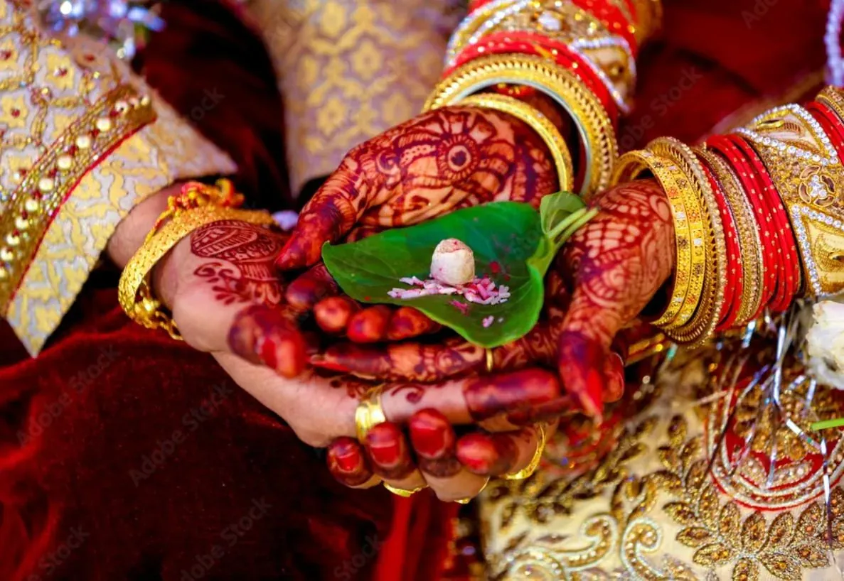 Hands adorned with henna and gold jewelry holding a betel leaf with a coconut during a traditional Indian wedding ceremony, showcasing cultural rituals and elegance.