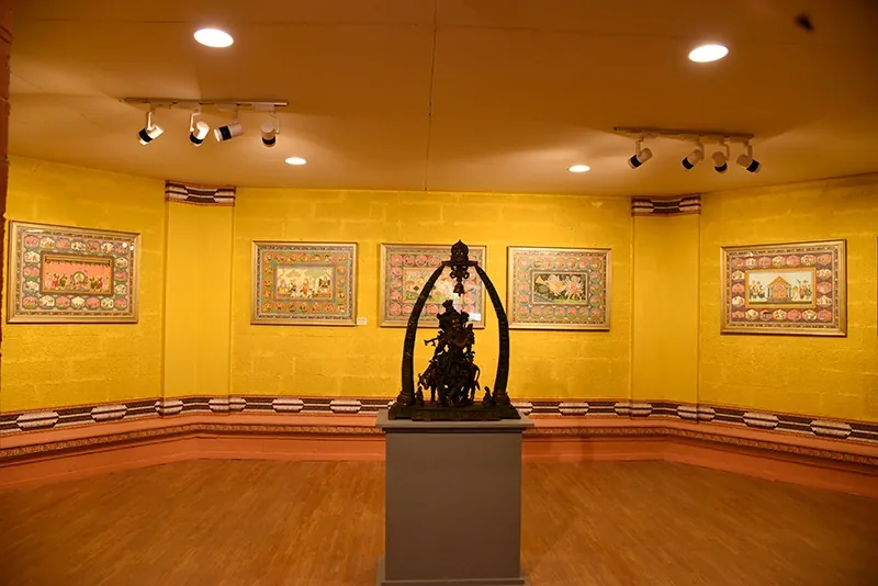 Interior view of a museum exhibit featuring a central dark sculpture on a pedestal, surrounded by colorful traditional paintings on yellow walls, illuminated by ceiling spotlights.
