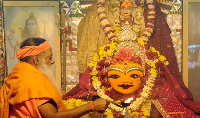 A devotee in orange robes offering prayers with a lit lamp to a richly adorned Hindu goddess statue, surrounded by flowers and garlands in a temple setting.
