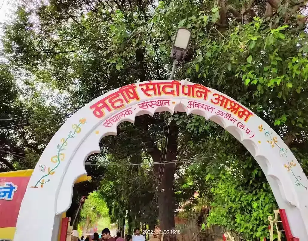 Entrance archway of Sandipani Ashram in Ujjain, featuring a vibrant sign with Hindi text, surrounded by lush green trees and a pathway, welcoming visitors to the historic site.