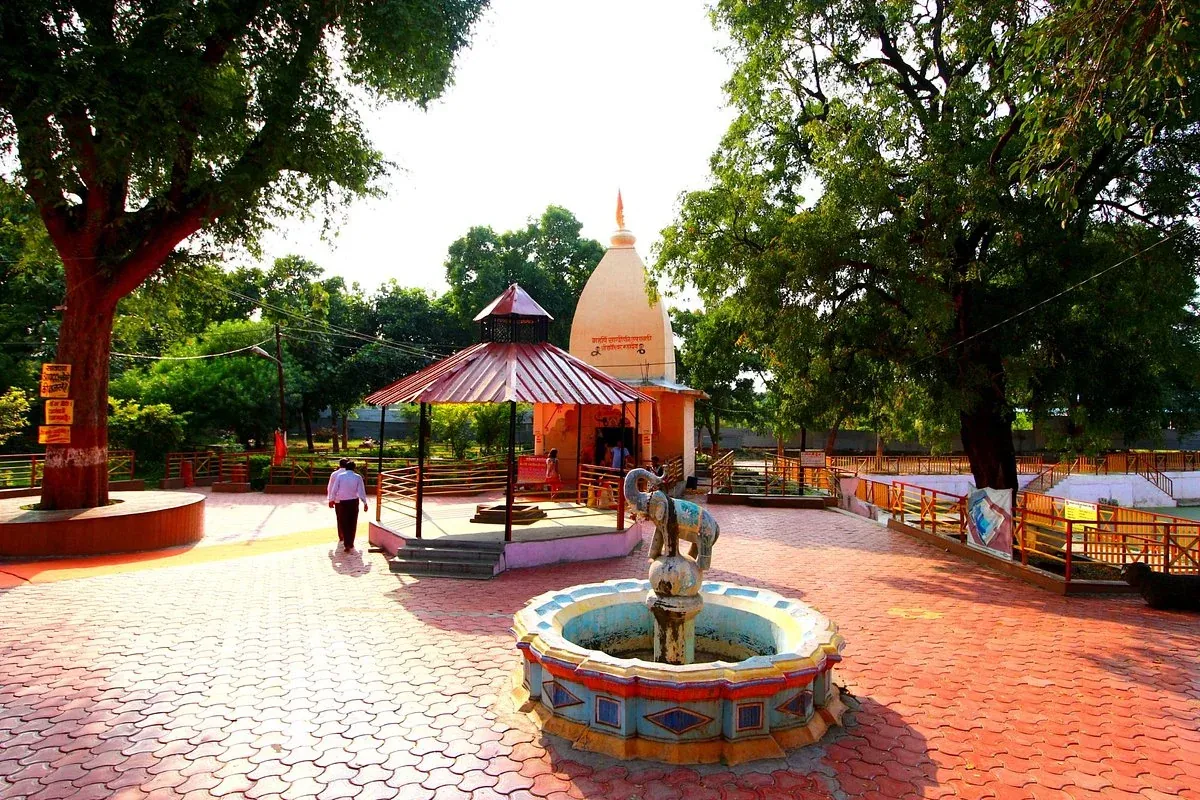 Scenic view of Sandipani Ashram featuring a traditional temple with a white spire, surrounded by lush green trees, a decorative fountain with elephant statues, and a paved walkway with a lone visitor.