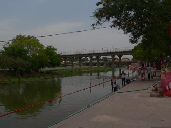 Serene riverside scene at a waterway with a bridge overpass, featuring a concrete walkway crowded with people, surrounded by trees and greenery, with anglers fishing along the calm waterfront during daytime.
