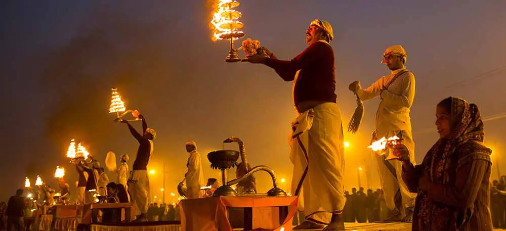 Priests performing Ganga Aarti with large oil lamps at a ghat during dusk, surrounded by devotees and glowing firelight.